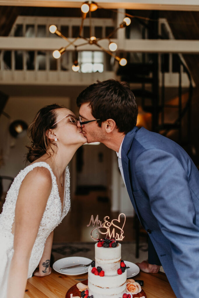 Bride and groom kiss over their wedding cake at a cozy Airbnb elopement, decorated with fresh berries and a “Mr. & Mrs.” topper