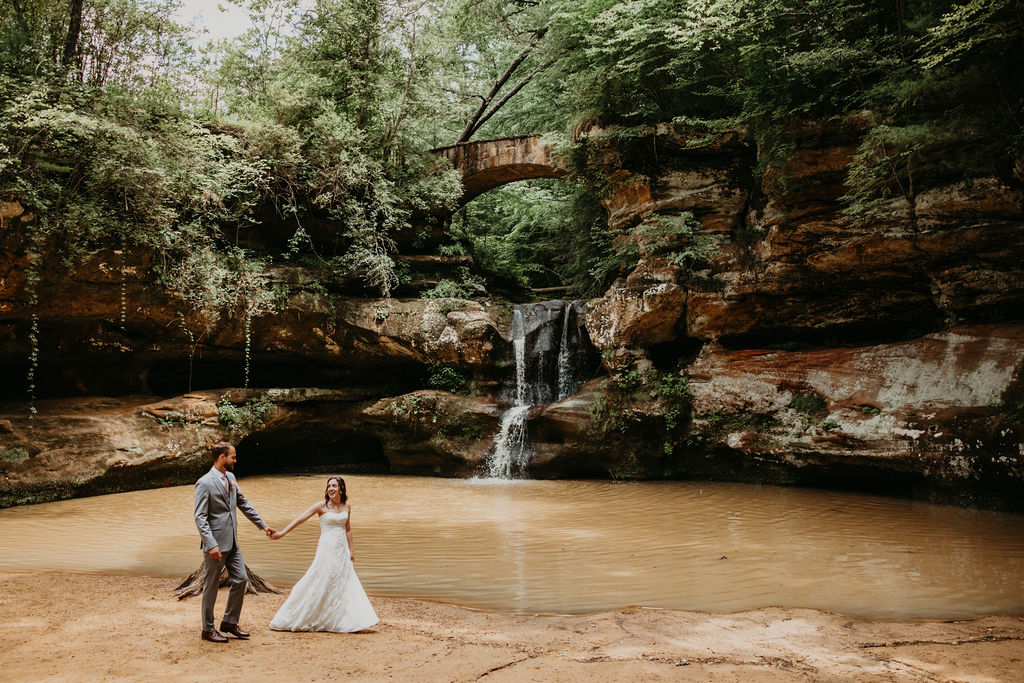 Bride and groom holding hands in front of a waterfall and natural stone bridge at Hocking Hills during their adventurous outdoor wedding session