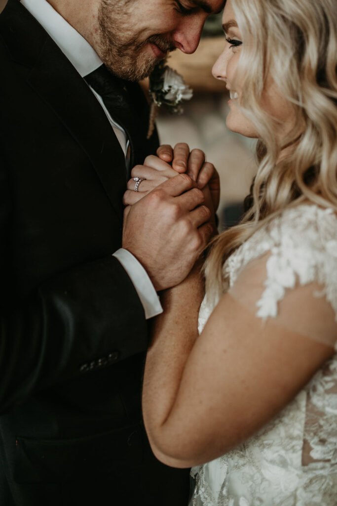 Close-up of bride and groom holding hands, smiling softly, with her wedding ring in focus and delicate lace details on her sleeve