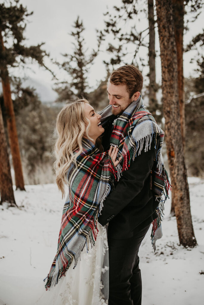 Bride and groom wrapped in a plaid blanket, laughing together in a snowy forest setting during a winter couple session