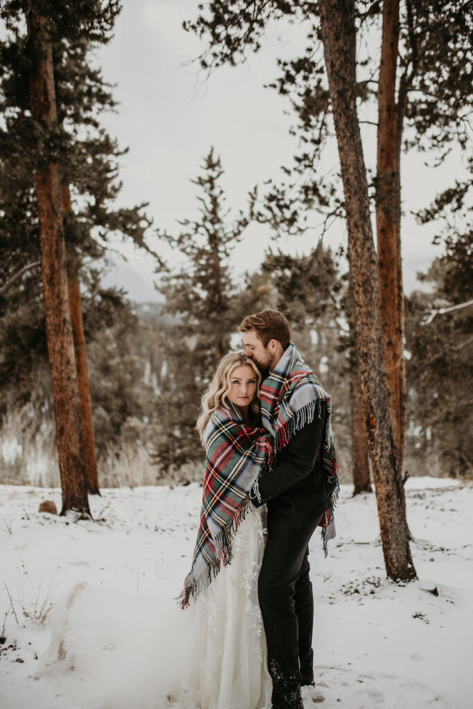 Adventurous couple wrapped in a plaid blanket in a snowy forest, bride looking at camera as groom kisses her head during a Norway couple photoshoot